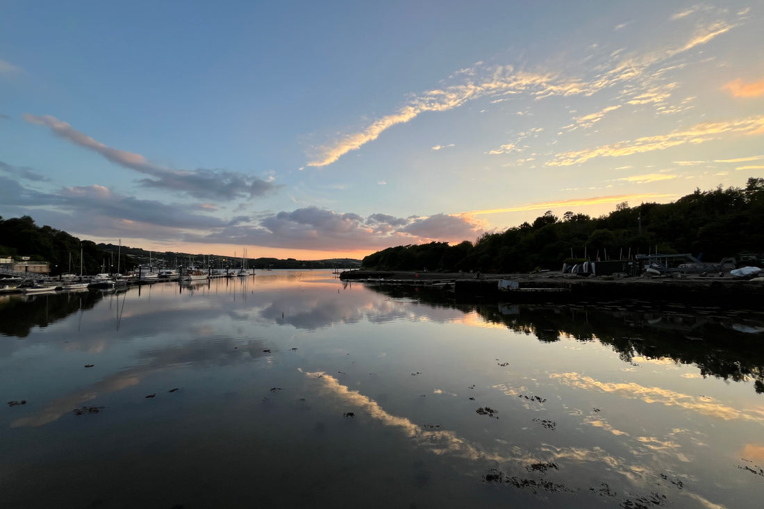 A picture of a creek at sunset, the clouds are reflected in the water. There are sailing bots moored up